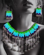 A woman wearing the Waterfall Boho Jewelry Set, with the vibrant enamel necklace resting along her neckline. The bold green, turquoise, and navy segments with glittery zigzag accents, paired with cascading silver-toned chains and beads, create a striking contrast against her outfit, showcasing the statement design's artistic and bohemian appeal — Muselot.