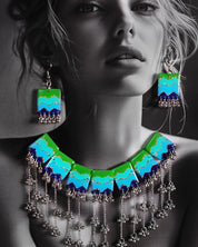 A model adorned in the Boho Jewelry Set, featuring a bold necklace with layered green, aqua, and deep blue panels highlighted with glitter accents. Silver-toned dangling chains and clustered beads sway below the enamel plates, adding movement and charm to the piece — Muselot.