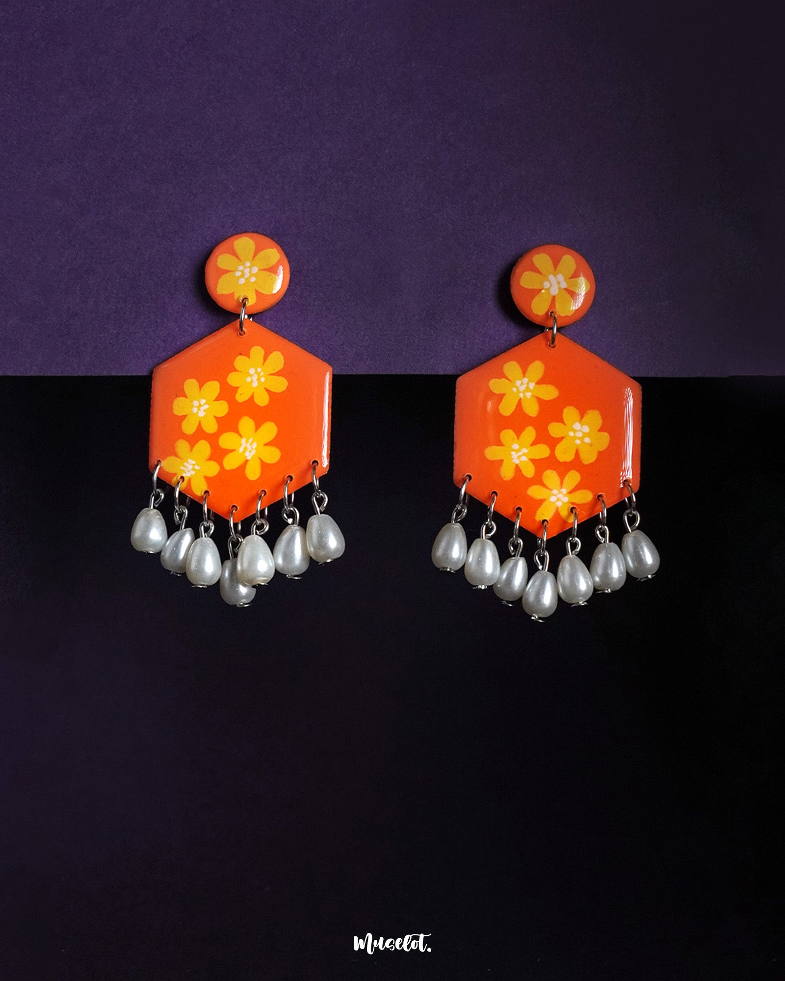 Zafraan floral earrings featuring round orange studs with yellow flower motifs, attached to hexagonal orange drops adorned with matching floral designs and finished with dangling white pearl-like beads, beautifully displayed against a deep purple background – Muselot.