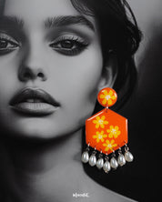 Close-up of model wearing the Zafraan floral earrings, showcasing round orange studs with yellow flower motifs attached to hexagonal orange drops with matching floral designs, accented by dangling white pearl-like beads, capturing their bright colors and intricate detailing – Muselot.