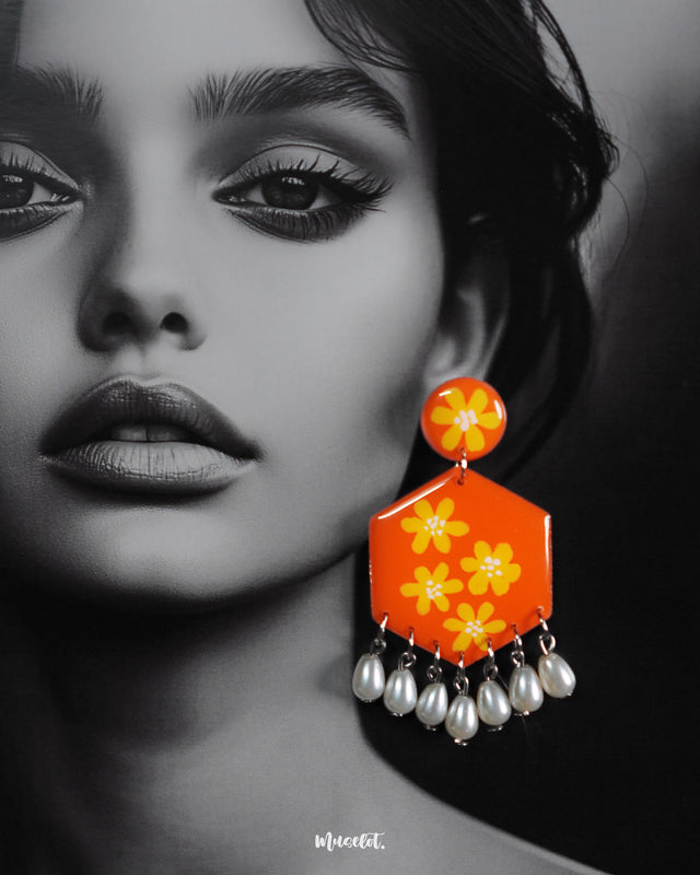 Close-up of model wearing the Zafraan floral earrings, showcasing round orange studs with yellow flower motifs attached to hexagonal orange drops with matching floral designs, accented by dangling white pearl-like beads, capturing their bright colors and intricate detailing – Muselot.