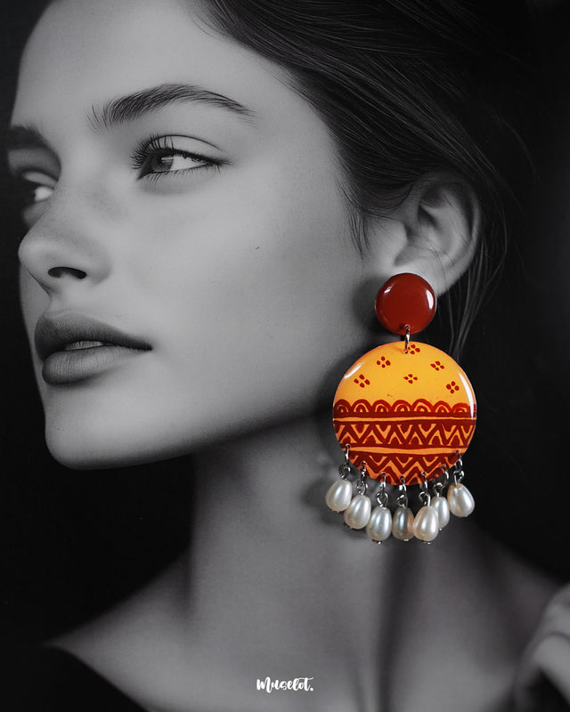 A closeup shot of a woman wears the Zardozi drop earrings, with warm mustard and rust-toned circular tops adorned in traditional hand-painted motifs and white pearl danglers—highlighting the rich craftsmanship and statement appeal of this artisanal pair — Muselot.