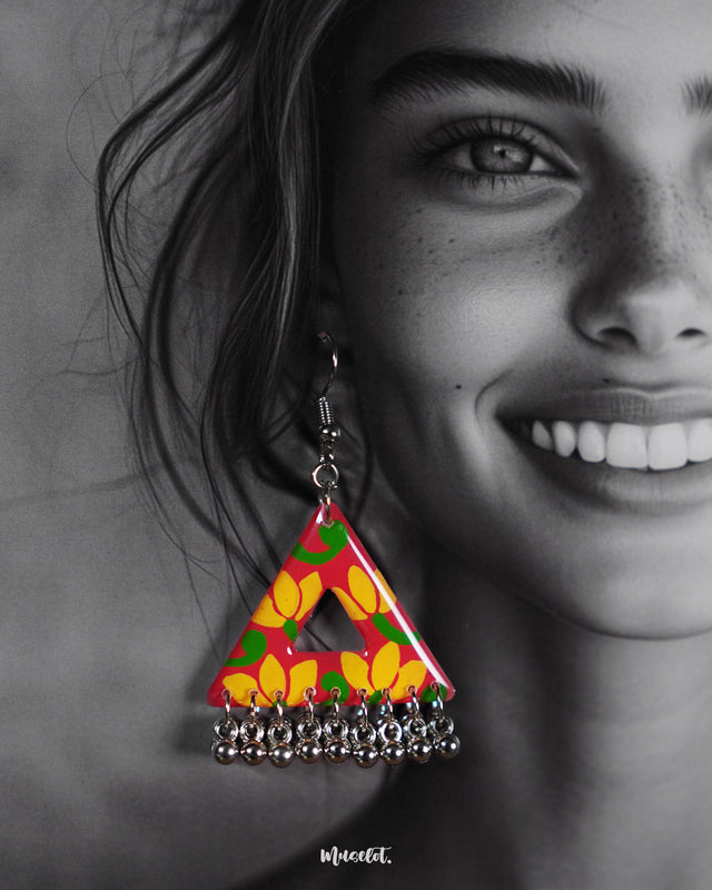 A close-up of a model wearing the Zarmin floral triangle earrings, highlighting the vivid pink and yellow floral hand-painted design and the row of silver ghungroos,showcasing their festive movement and handcrafted vibrance against the skin — Muselot.
