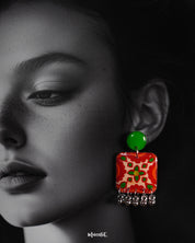 A close-up of a woman wearing the Zarnigar stud earrings, highlighting the striking hand-painted red and green floral design. The gleaming silver ghungroo drops sway delicately beneath the square motif, drawing attention to the vibrant detailing and folk-inspired artistry — Muselot.