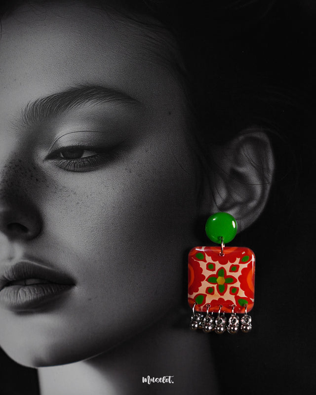 A close-up of a woman wearing the Zarnigar stud earrings, highlighting the striking hand-painted red and green floral design. The gleaming silver ghungroo drops sway delicately beneath the square motif, drawing attention to the vibrant detailing and folk-inspired artistry — Muselot.