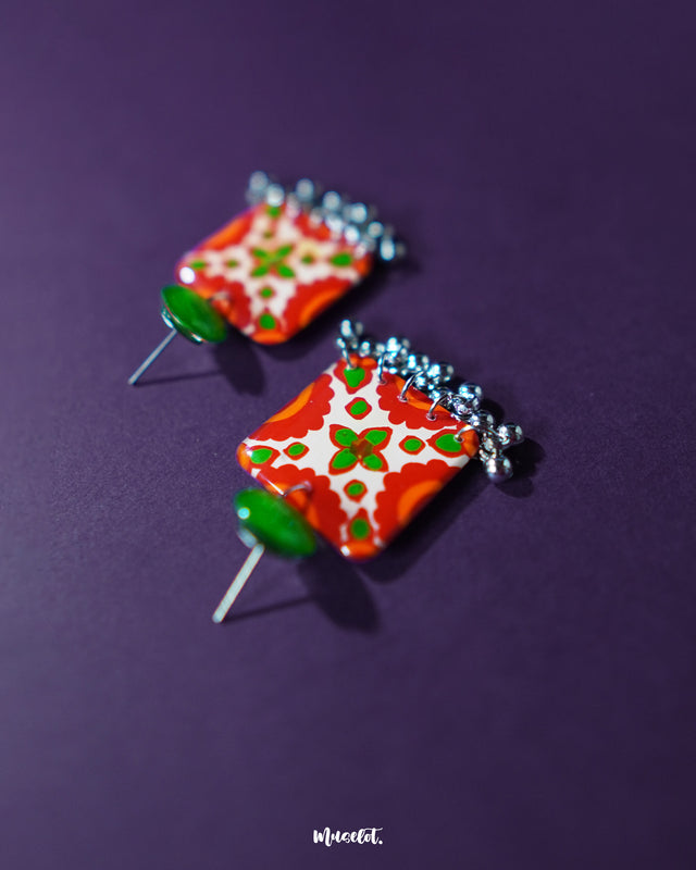 Zarnigar stud earrings set against a deep purple background, showcasing the vivid contrast of the hand-painted red and green floral pattern. The silver ghungroo drops add movement and charm to the bold square design, making the pair stand out with a festive yet elegant appeal — Muselot.