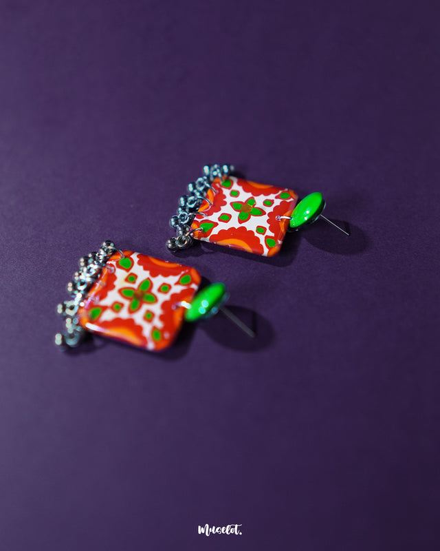 The Zarnigar stud earrings are showcased resting on a richly embroidered textile, their floral enamel work glowing in red, green, and ivory tones. The delicate silver ghungroos below add movement and a hint of melody, infusing the earrings with ethnic elegance — Muselot.