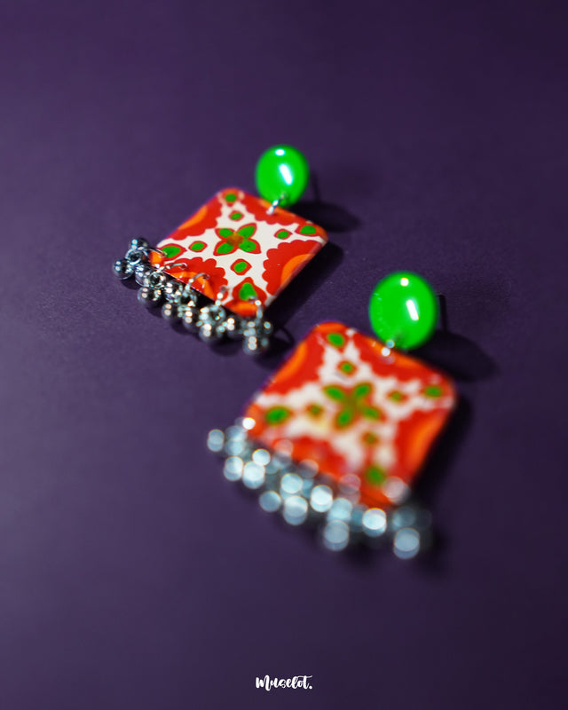 A close-up of the Zarnigar stud earrings highlighting their glossy finish and detailed hand-painted floral motif in red, green, and cream. The dangling silver ghungroos enhance the traditional charm, making these earrings a vibrant choice for festive and everyday looks — Muselot.