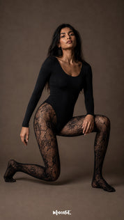 A woman in a black bodysuit kneeling while wearing full-length black designer lace stockings with an intricate rose floral fishnet pattern and closed feet-Muselot