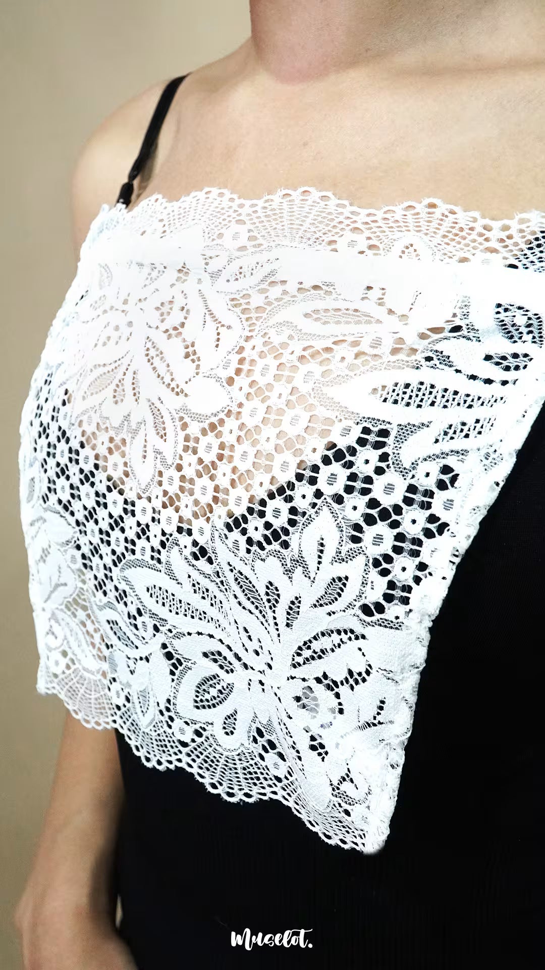 Side-angled close up of white lace camisole insert worn over black dress on beige background, showing broad lace design created to hide low necklines smoothly – muselot