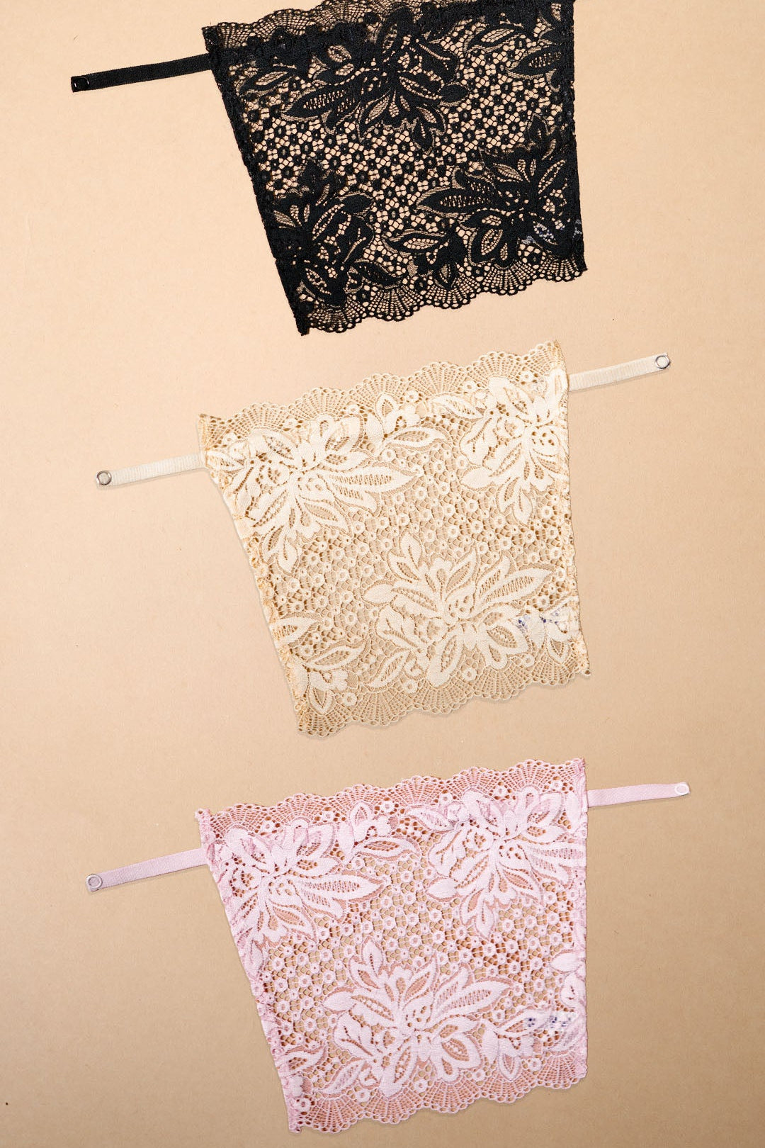 Three lace clip-on neckline covers in black, beige and light pink displayed on a beige background, featuring delicate floral lace detailing and adjustable straps. Designed to provide elegant coverage for deep neck tops, dresses and blouses, these lightweight mock camisole inserts add a feminine layered look while helping customize neckline coverage with comfort and style – Muselot