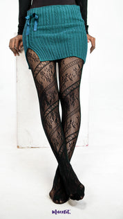 A model wearing sheer black lace stockings with floral fishnet design on a white background, styled with crossed legs - Muselot