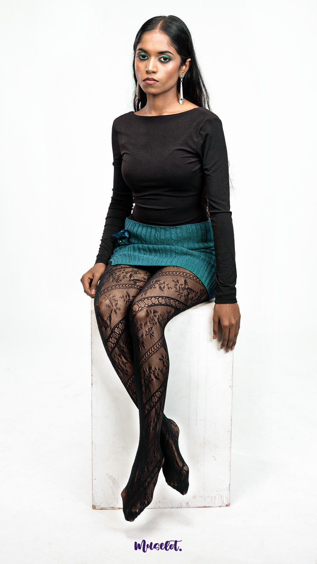 A model wearing black lace pantyhose sitting on a white block against a white background, showcasing stretch fit and detailed thigh pattern - Muselot