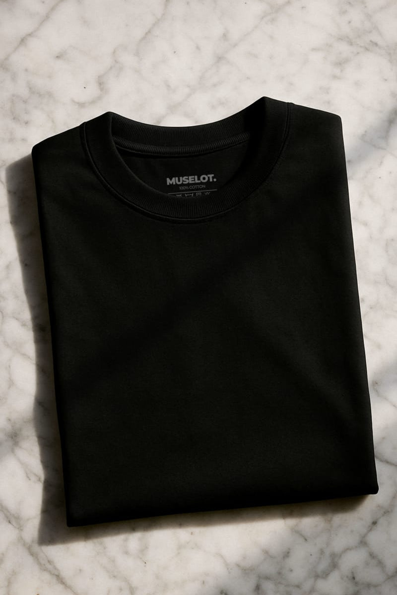 A unisex oversized black T-shirt is kept neatly folded on a sunlit marble surface, showcasing it's smooth texture and deep hue of the fabric, making it a bold yet minimal statement — Muselot.