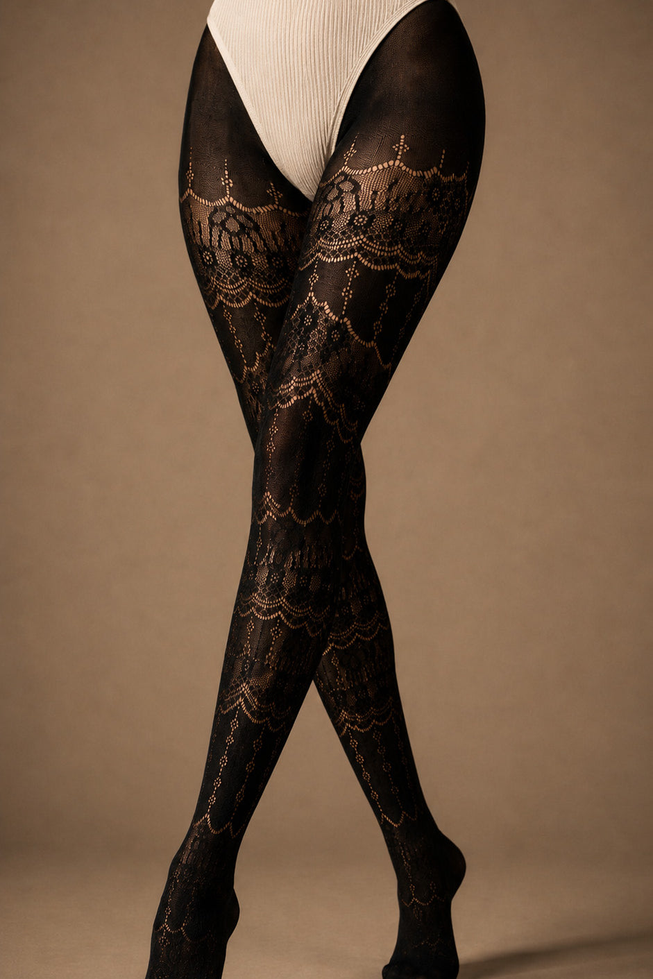 A focused view of crossed legs wearing full-length black lace stockings with intricate ornate motifs, semi-sheer sections and covered toes - Muselot