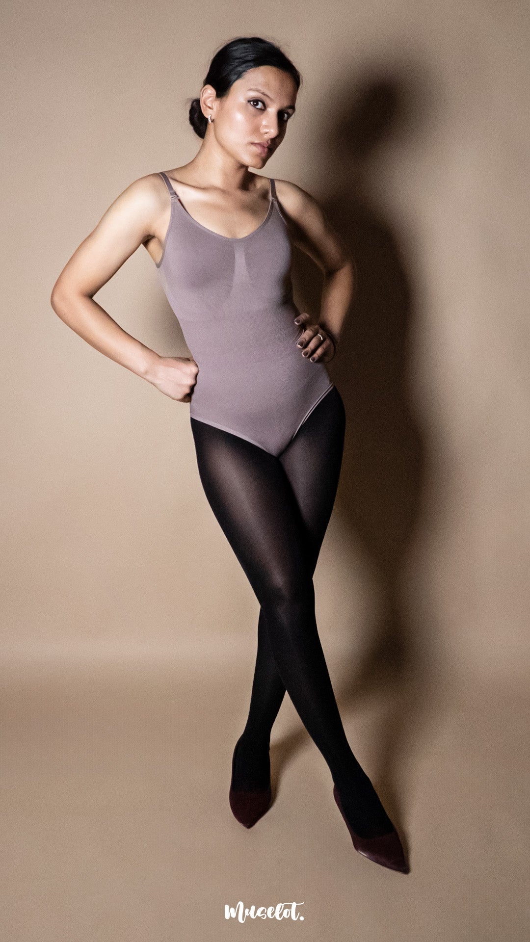 A model wearing black semi sheer tights styled with a bodysuit on a warm beige background, posing sideways to highlight smooth silhouette and no bunching fit – Muselot