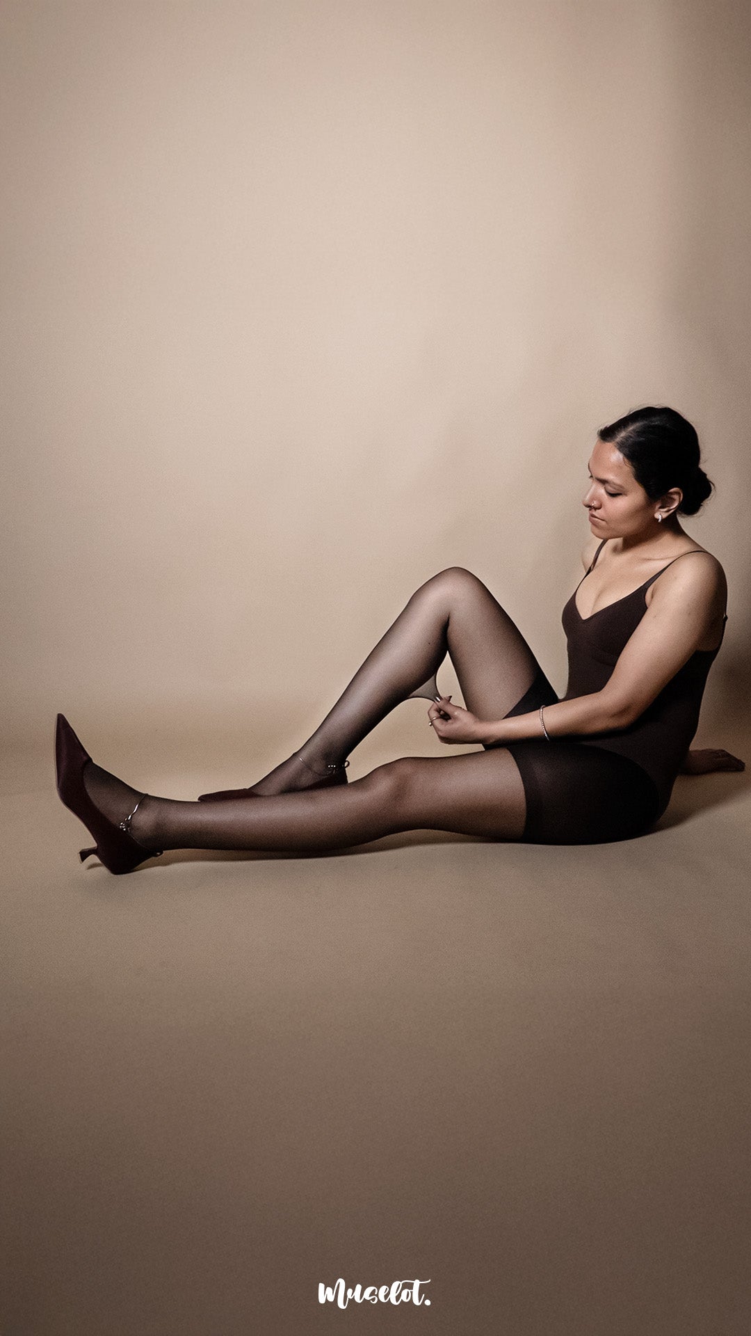 A model wearing transparent black pantyhose styled with heels, seated pose on a beige studio background highlighting flexibility and second-skin appearance — Muselot