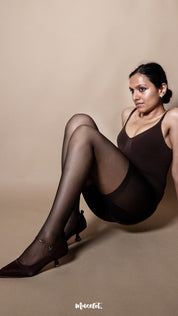 A model wearing ultra thin black pantyhose styled with heels, relaxed recline pose on a beige background showcasing lightweight feel and smooth finish — Muselot