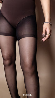 A model wearing ultra sheer black stockings styled with a fitted brown bodysuit, close-up thigh shot on a warm beige studio background highlighting transparent pantyhose finish and smooth coverage — Muselot