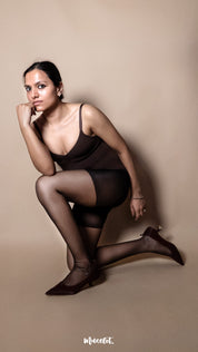 A model wearing black ultra sheer stockings styled with a bodysuit on a beige background demonstrating elasticity and tear-resistant fabric — Muselot